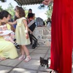 κόκκινο φόρεμα στα δεξιά της φωτογραφίας, ένα γατάκι στα πόδια της γυναίκας με το κόκκινο φόρεμα, δίπλα ακριβώς η θεία γονατιστή έχει στα χέρια της τη μικρή.