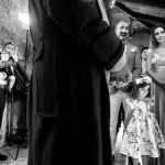 ασπρόμαυρη φωτογραφία από τη βάπτιση, στη μέση της φωτογραφίας το ράσο του παπά, δεξιά το ζεύγος με τη μεγάλη τους κόρη και αριστερά ένας μεσήλικας με το εγγόνι του αγκαλιά