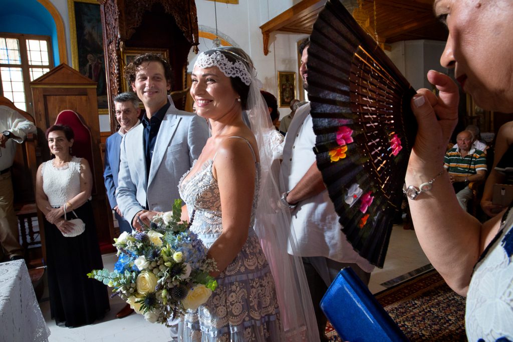 ο γαμπρός και η νύφη χαμογελούν καθώς ο πατέρας της την βοηθάει να δροσιστεί με μια βεντάλια πολύχρωμη.