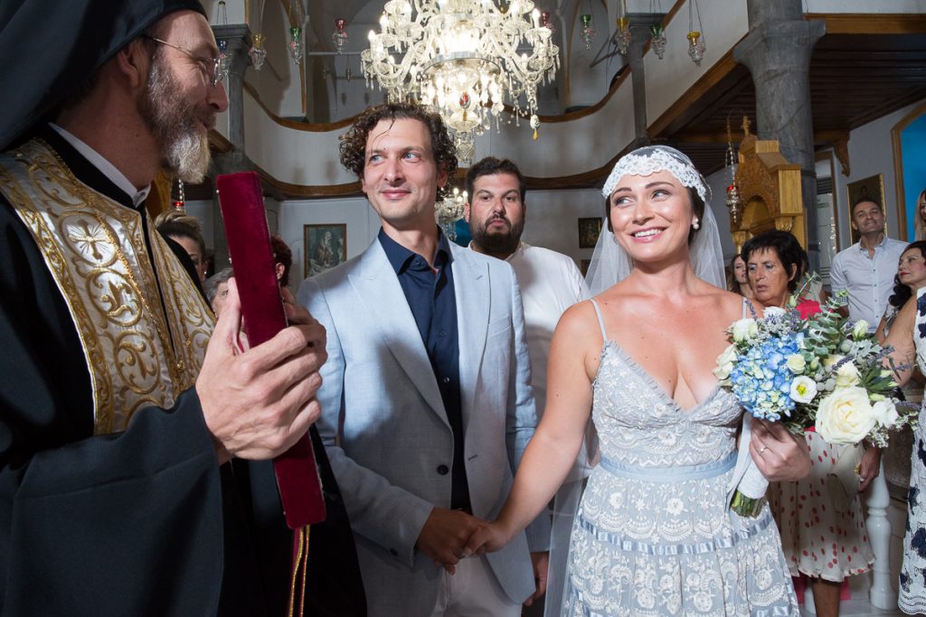 το ζευγάρι χαμογελαστό κοιτάει προς τη μεριά του ιερέα καθώς αυτός έχει στα χέρια του το ευαγγέλιο