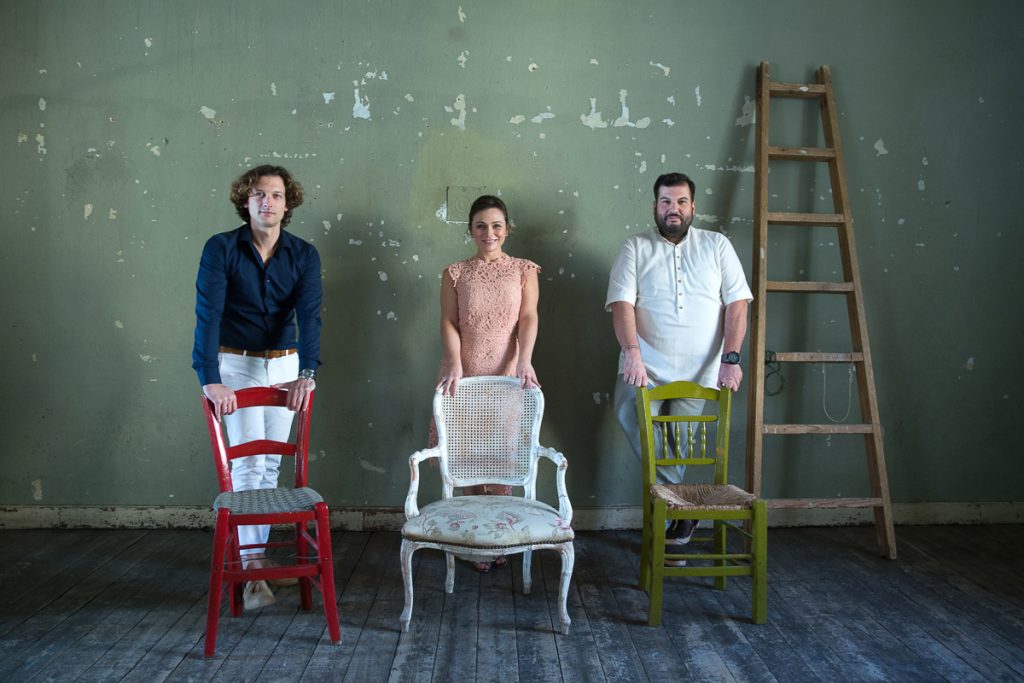 γαμπρός νύφη και κουμπάρος όρθιοι πίσω από τρεις καρέκλες, ακουμπάνε τα δυο τους χέρια σε αυτές.