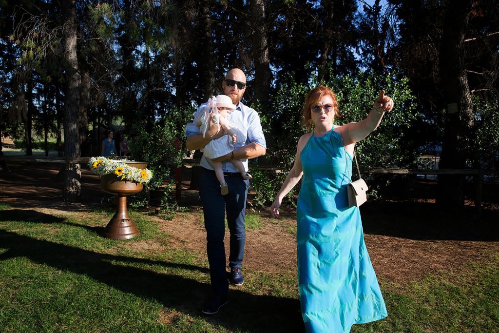 η γιαγιά δείχνει με απλωμένο το χέρι, δίπλα της ο γιος της με την ανιψιά του στα χέρια. πίσω από αυτούς η κολυμπήθρα.