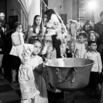 φωτογραφία ασπρόμαυρη από τη βάπτιση, Το κοριτσάκι με τη λαμπάδα και η νονά με το μωρό στην αγκαλιά κάνουν το γύρω της κολυμπήθρας.