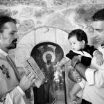 ο ιερές έχει ανοικτό το ευαγγέλιο και το στρέφει στο νονό για να διαβάσει το Πιστεύω ενώ κρατάει στα χέρια του το μικρό.